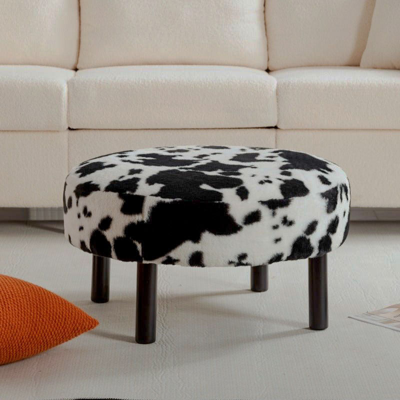 Round black-and-white cow-print upholstered ottoman with four black legs in front of a cream sofa