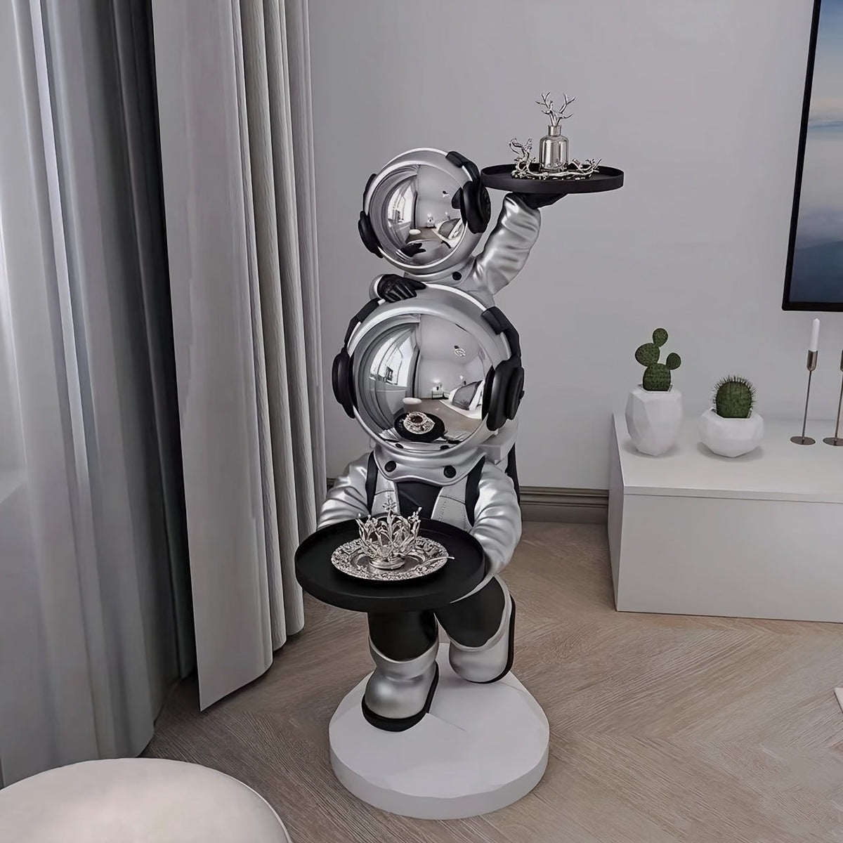 Silver astronaut side table with two black trays and mirrored visors beside gray curtains and a white cabinet