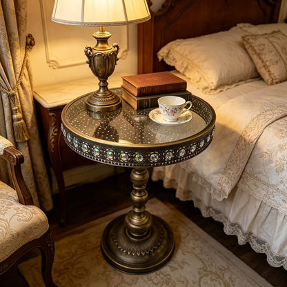 Bronze glass-top table used beside a bed and table lamp.