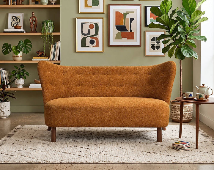 Caramel chenille loveseat with a curved button-tufted back and wood legs in a room with framed art and plants.