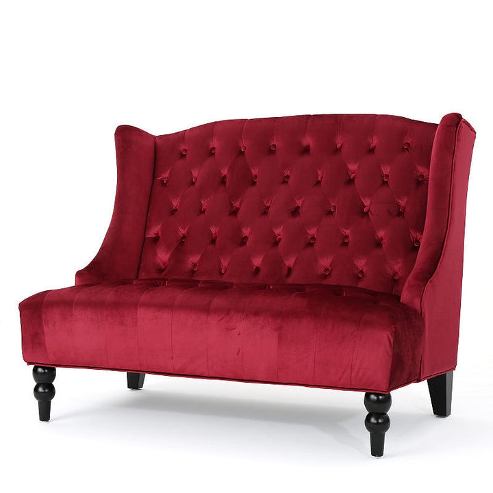 Angled front view of the wine red velvet loveseat showing its curved wing sides and button tufting.